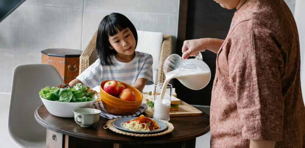 Crop faceless female standing near table and preparing healthy lunch for cute Asian kid at home