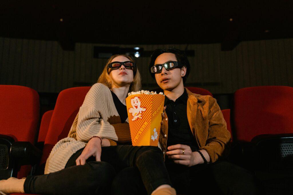 Two adults wearing 3D glasses watching a movie with popcorn in a cinema setting.