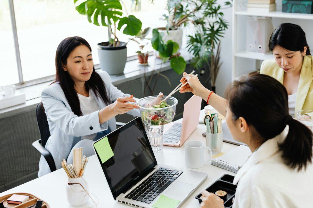 Three women sharing a healthy salad during a work break in a bright office setting.